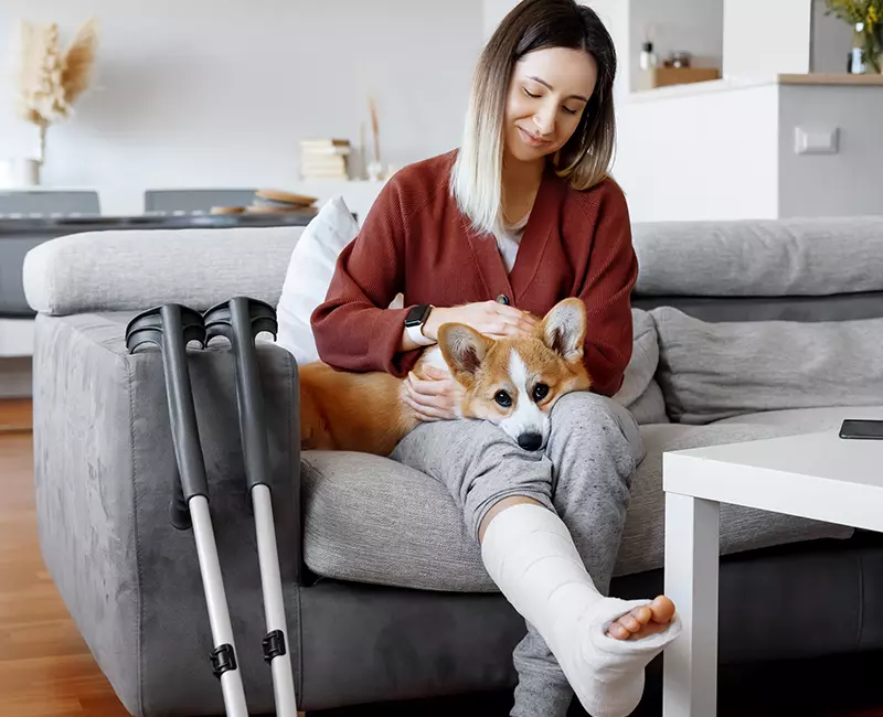 Femme dans son salon avec son pied dans le plâtre après accident sur son lieu de travail, son chien est sur ses genoux